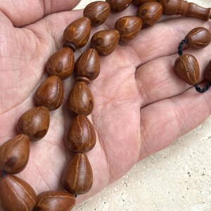 Puede incluir: Un collar de cuentas marrones en forma de l&aacute;grima, probablemente de madera, ensartadas en un cord&oacute;n negro. Las cuentas se sostienen en una mano, mostrando su veta natural de la madera y su textura suave. Las cuentas son de un tono c&aacute;lido y terroso.