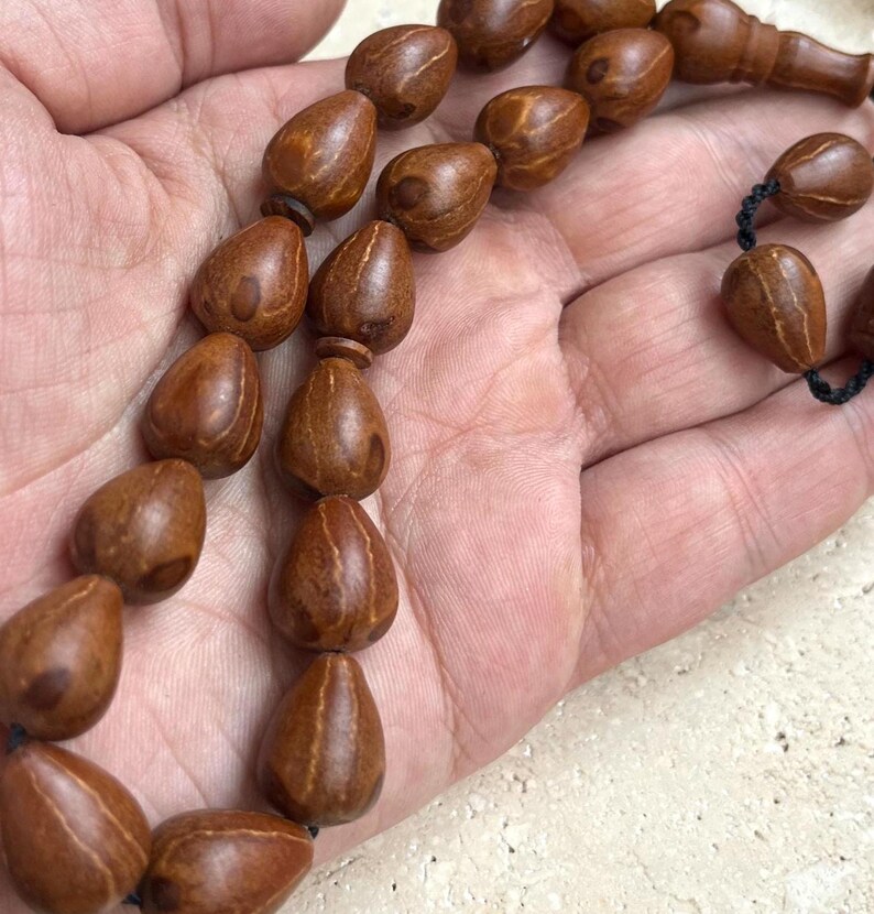 Puede incluir: Un collar de cuentas marrones en forma de l&aacute;grima, probablemente de madera, ensartadas en un cord&oacute;n negro. Las cuentas se sostienen en una mano, mostrando su veta natural de la madera y su textura suave. Las cuentas son de un tono c&aacute;lido y terroso.