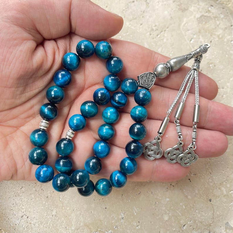Pode incluir: Um fio de contas de pedra olho de tigre azul. As contas redondas e polidas apresentam tons de azul escuro e claro. O fio &eacute; acentuado com componentes met&aacute;licos prateados e borlas.
