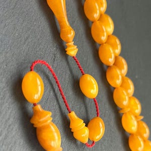 Puede incluir: Primer plano de un collar de cuentas de oraci&oacute;n. Las cuentas son de color &aacute;mbar c&aacute;lido, con forma ovalada. El collar tiene un cord&oacute;n rojo y piezas finales decorativas de color &aacute;mbar. El fondo es gris oscuro.