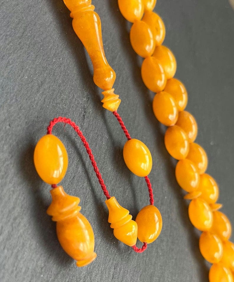Puede incluir: Primer plano de un collar de cuentas de oraci&oacute;n. Las cuentas son de color &aacute;mbar c&aacute;lido, con forma ovalada. El collar tiene un cord&oacute;n rojo y piezas finales decorativas de color &aacute;mbar. El fondo es gris oscuro.