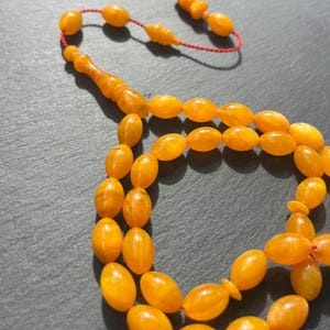 Puede incluir: Primer plano de un collar de cuentas de oraci&oacute;n de color &aacute;mbar. Las cuentas ovaladas est&aacute;n ensartadas en un cord&oacute;n rojo. Las cuentas brillantes est&aacute;n dispuestas en un patr&oacute;n circular. La borla tambi&eacute;n es de color &aacute;mbar.