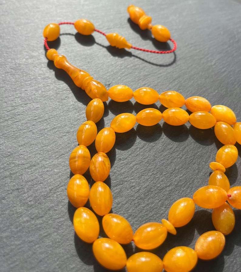 Puede incluir: Primer plano de un collar de cuentas de oraci&oacute;n de color &aacute;mbar. Las cuentas ovaladas est&aacute;n ensartadas en un cord&oacute;n rojo. Las cuentas brillantes est&aacute;n dispuestas en un patr&oacute;n circular. La borla tambi&eacute;n es de color &aacute;mbar.