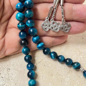 Pode incluir: Um fio de contas de pedra preciosa olho de tigre azul com detalhes prateados. As contas s&atilde;o de uma cor verde-azulado profundo com padr&otilde;es em espiral. O fio tem um pingente prateado decorativo e tr&ecirc;s pingentes prateados.