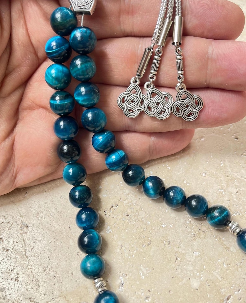 Pode incluir: Um fio de contas de pedra preciosa olho de tigre azul com detalhes prateados. As contas s&atilde;o de uma cor verde-azulado profundo com padr&otilde;es em espiral. O fio tem um pingente prateado decorativo e tr&ecirc;s pingentes prateados.
