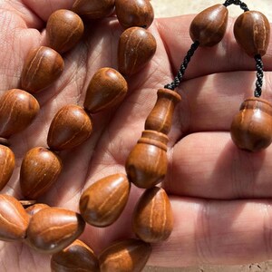 Puede incluir: Primer plano de un juego de cuentas de oraci&oacute;n de madera. Las cuentas tienen forma de l&aacute;grima y est&aacute;n ensartadas con un cord&oacute;n negro. La madera tiene un tono marr&oacute;n c&aacute;lido con vetas visibles. El juego incluye una pieza decorativa de madera.