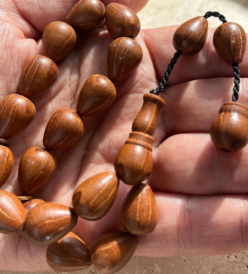 Puede incluir: Primer plano de un juego de cuentas de oraci&oacute;n de madera. Las cuentas tienen forma de l&aacute;grima y est&aacute;n ensartadas con un cord&oacute;n negro. La madera tiene un tono marr&oacute;n c&aacute;lido con vetas visibles. El juego incluye una pieza decorativa de madera.