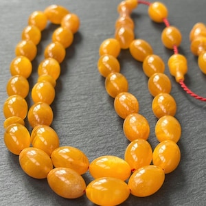 Puede incluir: Primer plano de un collar de cuentas de oraci&oacute;n de color &aacute;mbar. Las cuentas tienen forma ovalada y est&aacute;n ensartadas con un cord&oacute;n rojo. Las cuentas tienen un acabado brillante y est&aacute;n dispuestas sobre una superficie gris oscuro.