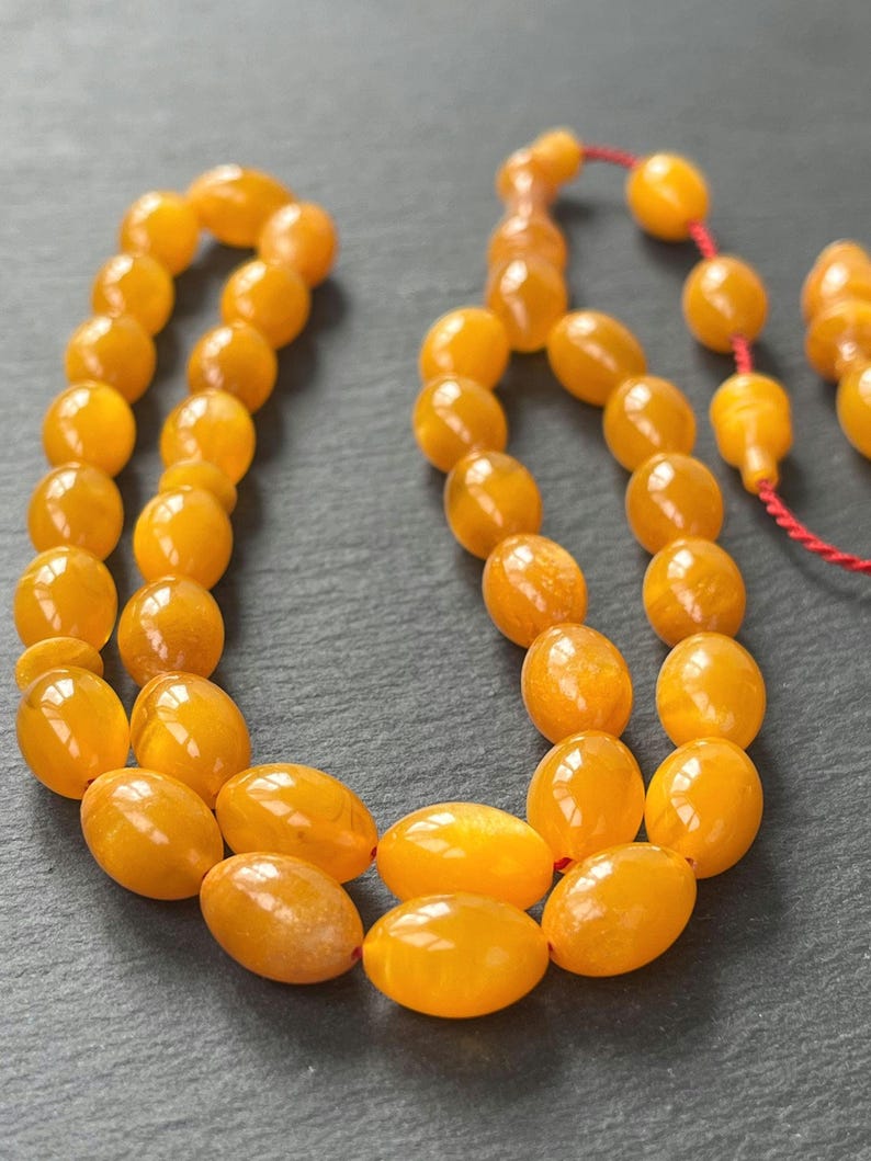 Puede incluir: Primer plano de un collar de cuentas de oraci&oacute;n de color &aacute;mbar. Las cuentas tienen forma ovalada y est&aacute;n ensartadas con un cord&oacute;n rojo. Las cuentas tienen un acabado brillante y est&aacute;n dispuestas sobre una superficie gris oscuro.