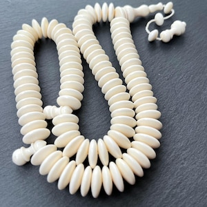 May include: Cream-coloured prayer beads with oval beads and a decorative tassel. The beads are strung together, forming a long, looping strand. The beads are arranged in a circular pattern.