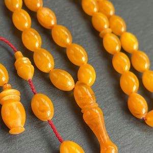 Puede incluir: Primer plano de un collar de cuentas de oraci&oacute;n de color &aacute;mbar. Las cuentas son de forma ovalada y est&aacute;n ensartadas en un cord&oacute;n rojo. El punto focal son las cuentas, con una pieza final decorativa y un elemento similar a una borla. El fondo es una superficie gris oscuro.