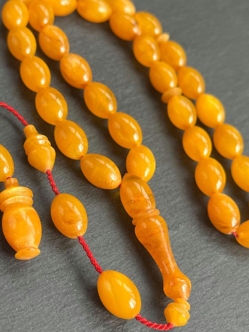 Puede incluir: Primer plano de un collar de cuentas de oraci&oacute;n de color &aacute;mbar. Las cuentas son de forma ovalada y est&aacute;n ensartadas en un cord&oacute;n rojo. El punto focal son las cuentas, con una pieza final decorativa y un elemento similar a una borla. El fondo es una superficie gris oscuro.