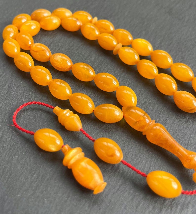 Puede incluir: Primer plano de cuentas de oraci&oacute;n de color &aacute;mbar con cuentas de forma ovalada ensartadas en un cord&oacute;n rojo. Las cuentas tienen un acabado pulido y brillante. La borla y la pieza final tambi&eacute;n son de color &aacute;mbar. El fondo es una superficie gris oscuro.