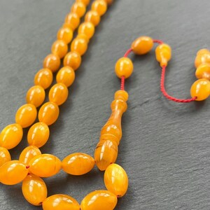 Puede incluir: Primer plano de un collar de cuentas de oraci&oacute;n. Las cuentas ovaladas, de color &aacute;mbar, est&aacute;n ensartadas en un cord&oacute;n rojo. La borla y la pieza final tambi&eacute;n son de color &aacute;mbar. El fondo es una superficie gris oscuro.