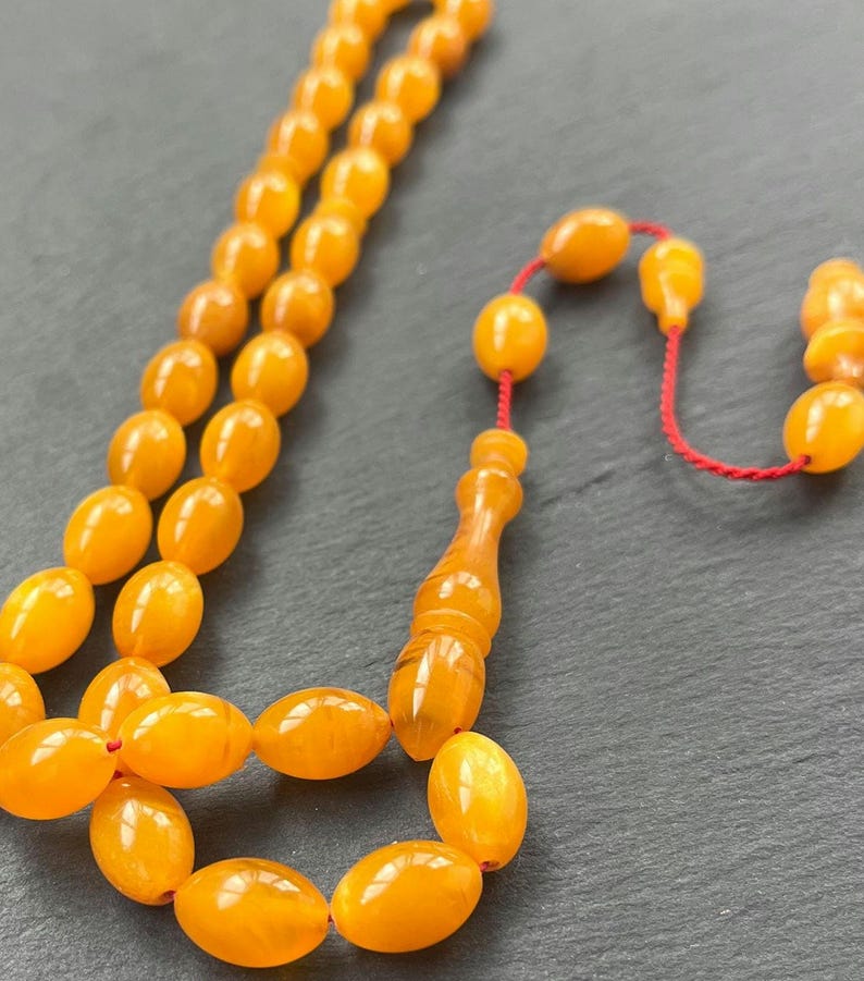 Puede incluir: Primer plano de un collar de cuentas de oraci&oacute;n. Las cuentas ovaladas, de color &aacute;mbar, est&aacute;n ensartadas en un cord&oacute;n rojo. La borla y la pieza final tambi&eacute;n son de color &aacute;mbar. El fondo es una superficie gris oscuro.