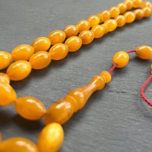 Puede incluir: Primer plano de un collar de cuentas de oraci&oacute;n de color &aacute;mbar. Las cuentas tienen forma ovalada y est&aacute;n ensartadas con un cord&oacute;n rojo. Las cuentas tienen un acabado brillante y est&aacute;n dispuestas sobre una superficie oscura.