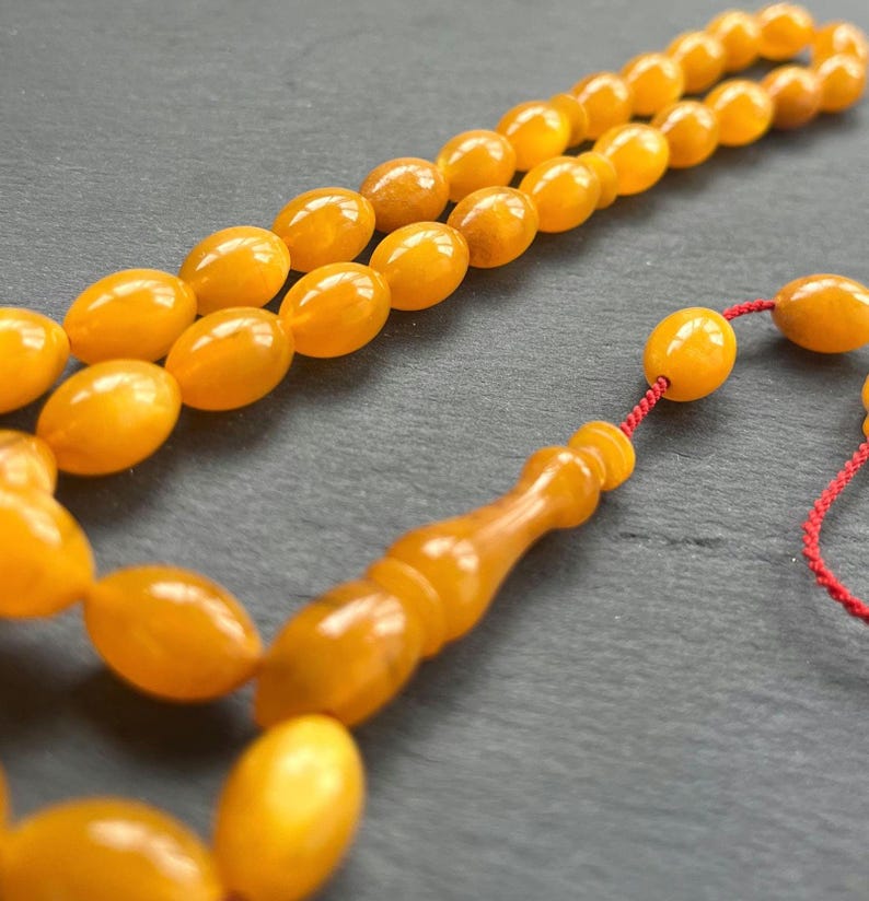 Puede incluir: Primer plano de un collar de cuentas de oraci&oacute;n de color &aacute;mbar. Las cuentas tienen forma ovalada y est&aacute;n ensartadas con un cord&oacute;n rojo. Las cuentas tienen un acabado brillante y est&aacute;n dispuestas sobre una superficie oscura.
