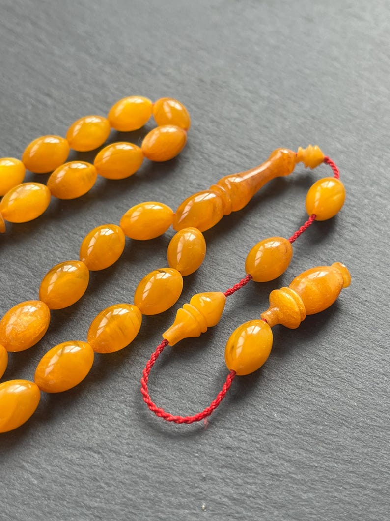 Puede incluir: Primer plano de un juego de cuentas de oraci&oacute;n. Las cuentas tienen forma ovalada y son de color &aacute;mbar, ensartadas con un cord&oacute;n rojo. El juego incluye una pieza decorativa y un extremo en forma de borla. El fondo es una superficie gris oscuro.