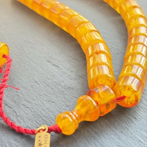 May include: Close-up of a strand of amber-coloured prayer beads. The beads are cylindrical with rounded edges and strung on a red cord. A small gold tag with the word "OMARI" is attached.