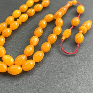 Puede incluir: Primer plano de un collar de cuentas de oraci&oacute;n de color &aacute;mbar. Las cuentas tienen forma ovalada y est&aacute;n ensartadas con un cord&oacute;n rojo. Las cuentas tienen un acabado brillante y est&aacute;n dispuestas sobre una superficie oscura.
