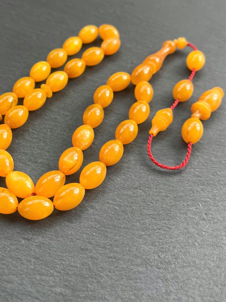 Puede incluir: Primer plano de un collar de cuentas de oraci&oacute;n de color &aacute;mbar. Las cuentas tienen forma ovalada y est&aacute;n ensartadas con un cord&oacute;n rojo. Las cuentas tienen un acabado brillante y est&aacute;n dispuestas sobre una superficie oscura.