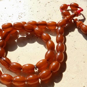 K&ouml;nnte beinhalten: Eine Kette aus bernsteinfarbenen Gebetsperlen mit oval geformten Perlen. Die Perlen sind aufgef&auml;delt, mit einem dekorativen Endst&uuml;ck und einer roten Quaste. Die Perlen sind auf einer hellen Oberfl&auml;che angeordnet.
