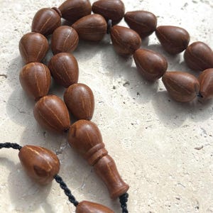 Puede incluir: Primer plano de un collar de cuentas de oraci&oacute;n. Las cuentas en forma de l&aacute;grima son de madera marr&oacute;n oscuro. El collar tiene un cord&oacute;n negro y una pieza decorativa de madera. Las cuentas est&aacute;n dispuestas en un patr&oacute;n circular.