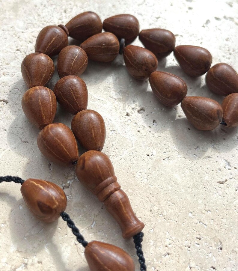 Puede incluir: Primer plano de un collar de cuentas de oraci&oacute;n. Las cuentas en forma de l&aacute;grima son de madera marr&oacute;n oscuro. El collar tiene un cord&oacute;n negro y una pieza decorativa de madera. Las cuentas est&aacute;n dispuestas en un patr&oacute;n circular.