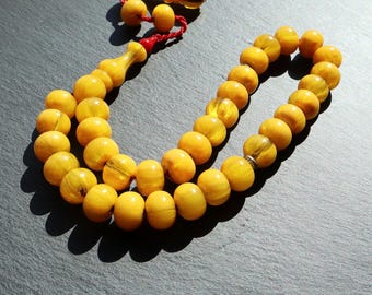 Pode incluir: Um fio de contas de oração amarelas com borla e peça final decorativa. As contas são de cor amarelo quente e parecem ser feitas de um material translúcido. As contas são enfiadas num cordão vermelho.