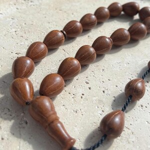 Puede incluir: Primer plano de un collar de cuentas de oraci&oacute;n. Las cuentas tienen forma de l&aacute;grima y est&aacute;n hechas de madera marr&oacute;n. Las cuentas est&aacute;n ensartadas con un cord&oacute;n negro. El collar descansa sobre una superficie clara y texturizada.