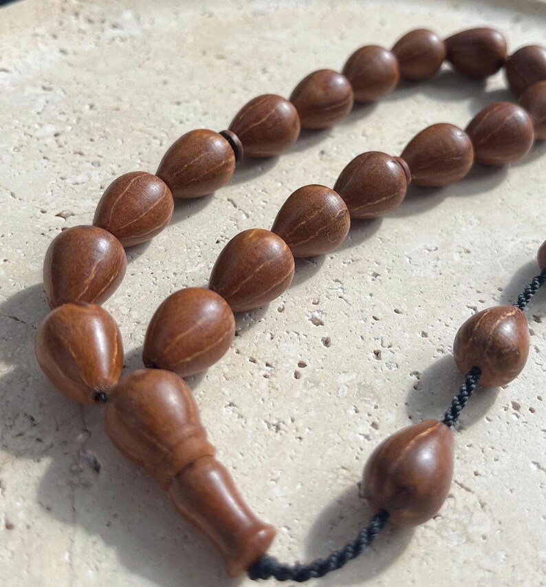 Puede incluir: Primer plano de un collar de cuentas de oraci&oacute;n. Las cuentas tienen forma de l&aacute;grima y est&aacute;n hechas de madera marr&oacute;n. Las cuentas est&aacute;n ensartadas con un cord&oacute;n negro. El collar descansa sobre una superficie clara y texturizada.