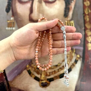 May include: A strand of peach-colored prayer beads with a decorative silver tassel. The beads are held in front of a painting of a person with a gold necklace and earrings.