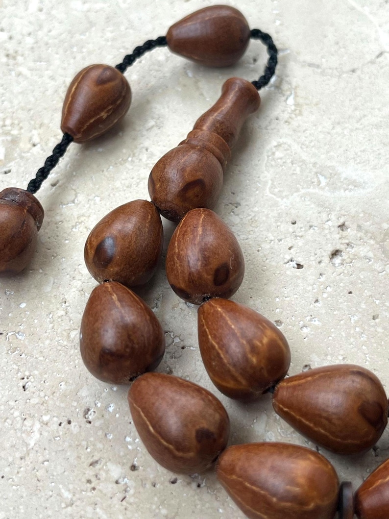 Puede incluir: Primer plano de un rosario de madera, con cuentas marrones en forma de l&aacute;grima ensartadas en un cord&oacute;n negro. Las cuentas tienen un acabado liso y pulido y est&aacute;n conectadas por una pieza decorativa de madera. El fondo es una superficie clara y texturizada.