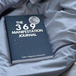 Może przedstawiać: Ciemnoniebieski dziennik zatytułowany "THE 369 MANIFESTATION JOURNAL" z grafiką księżyca i detalami gwiazd. Dziennik spoczywa na szarej narzucie. Tekst "Calm • Aligned • Intentional" znajduje się na dole.