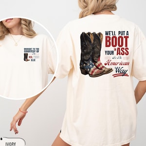 Puede incluir: Camiseta de color marfil con un diseño gráfico de botas de vaquero con la bandera americana. El texto en la parte posterior dice: "We'll put a boot in your ass, it's the American way." La parte delantera tiene un gráfico más pequeño y texto.