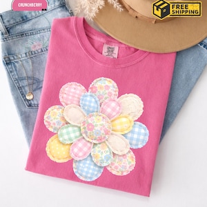 以下が含まれることがあります： パステルギンガムとフローラル柄の大きなアップリケの花柄が特徴のピンク色のComfort Colors Tシャツ。Tシャツは、青いジーンズとタンハットの上に置かれています。画像には「送料無料」のグラフィックも含まれています。