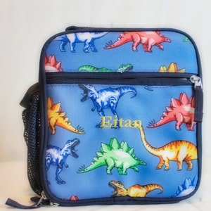 May include: Blue lunch bag with a dinosaur print and the name "Eitan" embroidered on the front. The bag has a zipper closure and a mesh side pocket.