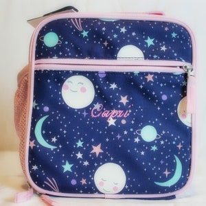 May include: Navy blue lunch bag with a celestial design featuring smiling moons, planets, stars, and the name "Capri" embroidered in pink. The bag has a pink zipper, trim, and side mesh pocket.