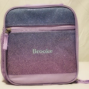 May include: Purple lunch bag with a zipper closure and a sparkly ombre design. The bag has a mesh side pocket and the name "Brooke" is embroidered on the front.