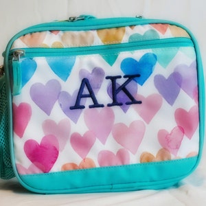 May include: A turquoise lunch bag with a white background and a pattern of watercolor hearts. The bag has a zipper closure and a mesh side pocket. The initials "AK" are embroidered on the front of the bag.