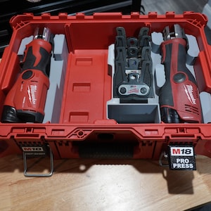 May include: A red Milwaukee M18 Pro Press tool case with a molded interior containing two red and black cordless power tools and a set of black metal press jaws. The case features M18 branding on the front latches and a Pro Press label.