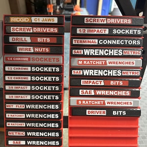 Puede incluir: Dos organizadores de herramientas rojos y negros con etiquetas para diversas herramientas, incluyendo "Destornilladores", "Brocas" y "Vasos". Las etiquetas están en texto blanco y rojo.