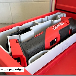 May include: A red and black Milwaukee oscillating multi-tool is stored in a red toolbox with gray compartments. The tool is labeled with the Milwaukee logo. The toolbox is open, revealing the tool and its storage.