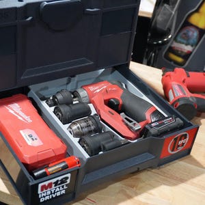 以下が含まれることがあります： 黒いTanos収納ケースの開いた引き出しの中に、赤いMilwaukee M12インストールドライバー、複数の交換可能なチャックアタッチメント、赤いアクセサリーケースが入っています。ケースの外側には赤いMilwaukeeの電動工具が置かれています。