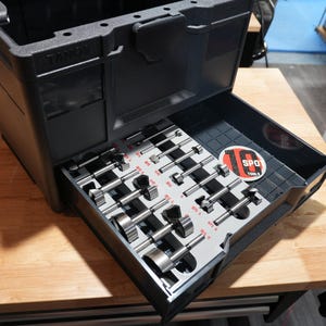 Può includere: Una valigetta in plastica nera con un cassetto aperto contenente un set di punte metalliche organizzate in un inserto in schiuma grigio chiaro. Il cassetto presenta un adesivo circolare con il testo 10 SPOT TOOL B e etichette rosse.
