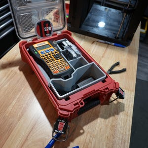 Può includere: Una custodia in plastica rossa con coperchio trasparente contenente un'etichettatrice portatile nera e arancione e divisori interni grigi. La custodia presenta un adesivo circolare con il testo 18 SPOT e due etichette nere con LABEL MAKER.