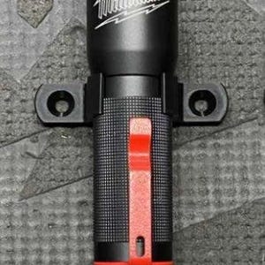 Pode incluir: Lanterna preta com o logotipo "Milwaukee" em branco. A lanterna tem um detalhe vermelho e uma pega texturizada. Está montada numa braçadeira preta com dois orifícios de montagem.