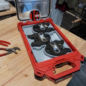 May include: A red and gray hard-shell carrying case containing three black metal crimping tool heads, with a circular label reading 10 SPOT TOOLS inside the lid. Two small pliers with red handles are positioned to the left of the open case.