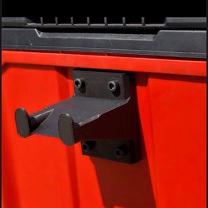 Op de afbeelding: Zwarte metalen gereedschapshouder gemonteerd op een rode ondergrond. De houder heeft twee gebogen armen die uitsteken vanaf een rechthoekige basis, vastgezet met schroeven. De achtergrond is een zwart oppervlak.