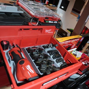 Puede incluir: Un sistema de almacenamiento de herramientas Milwaukee Packout rojo y negro con un cajón abierto que contiene una sierra recíproca inalámbrica roja y varios accesorios negros organizados en un inserto de plástico gris, con una caja Craftsman cerca.